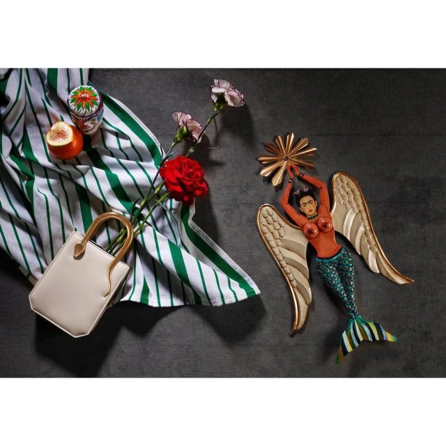 Still life composition features a mermaid figure, handbag, flowers, striped cloth, and fruit on a grey surface, creating a whimsical scene.