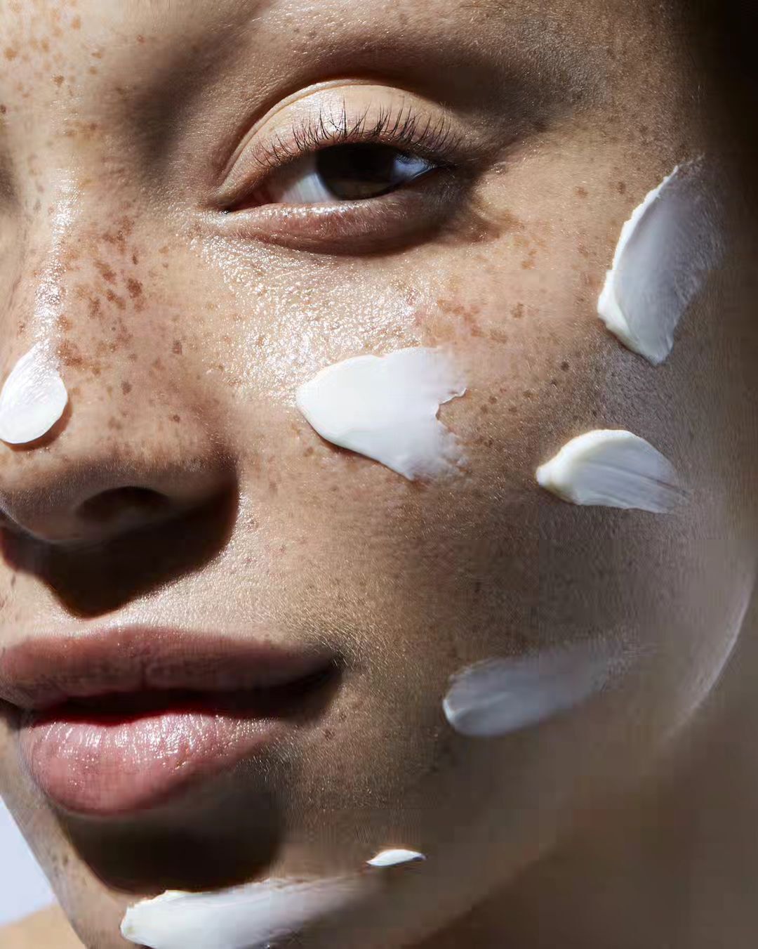 Close-up of a woman's face with fair skin and freckles, adorned with dabs of white cream, emphasizing skincare and beauty.