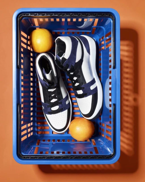 High-top sneakers in a blue shopping basket alongside two yellow lemons against an orange background creating a colorful product shot.