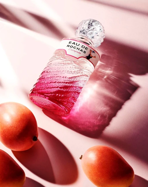 Eau de Rochas l'Escapade fragrance bottle on a pink surface with fresh mangoes, showcasing a beauty product with fruity elements.