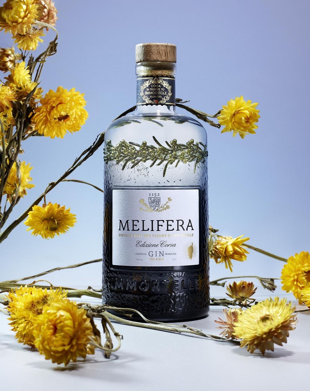 Melifera French Gin bottle surrounded by dried yellow flowers against a light blue background in a product shot.