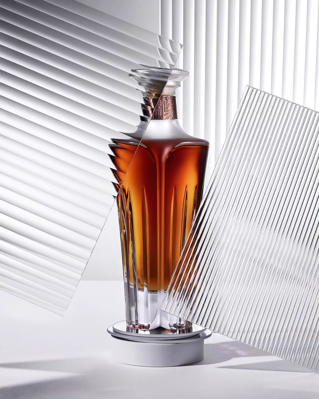 Crystal decanter with amber liquid stands on a white platform, textured glass panels, and venetian blind background add geometric detail.