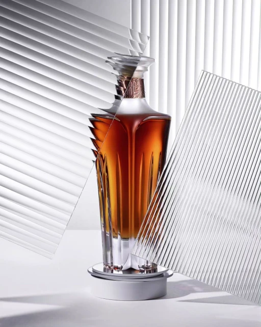 Crystal decanter with amber liquid stands on a white platform, textured glass panels, and venetian blind background add geometric detail.