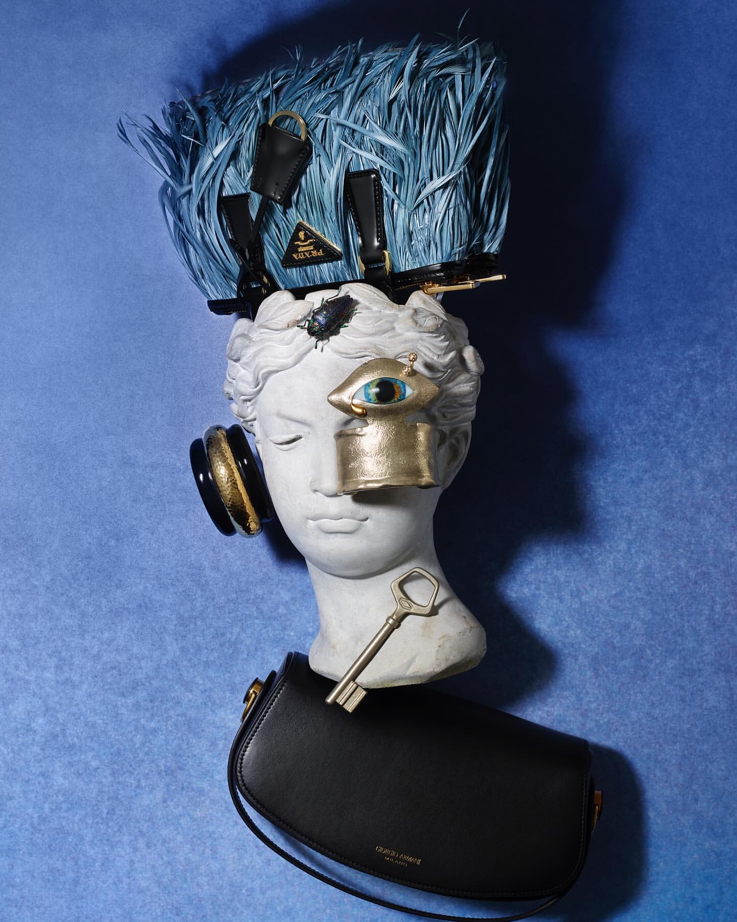 Still life: bust with headphones, blue feather hat, designer bag, and key against a textured blue background. Fashion and art concept.