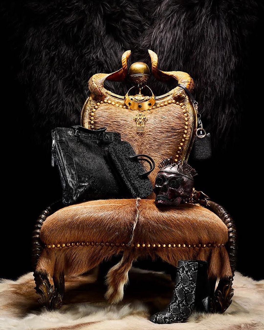 Ornate throne chair draped with fur and adorned with black handbags, skull mask, and snakeskin boots against a dark backdrop.