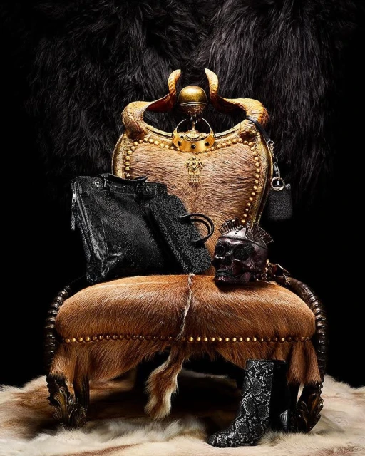 Ornate throne chair draped with fur and adorned with black handbags, skull mask, and snakeskin boots against a dark backdrop.