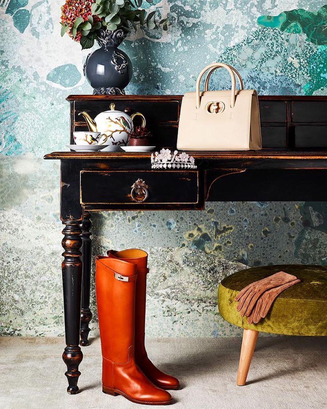 Still life of designer leather handbag, boots, tiara, vanity table, and floral arrangement in a studio setting with artistic wallpaper.