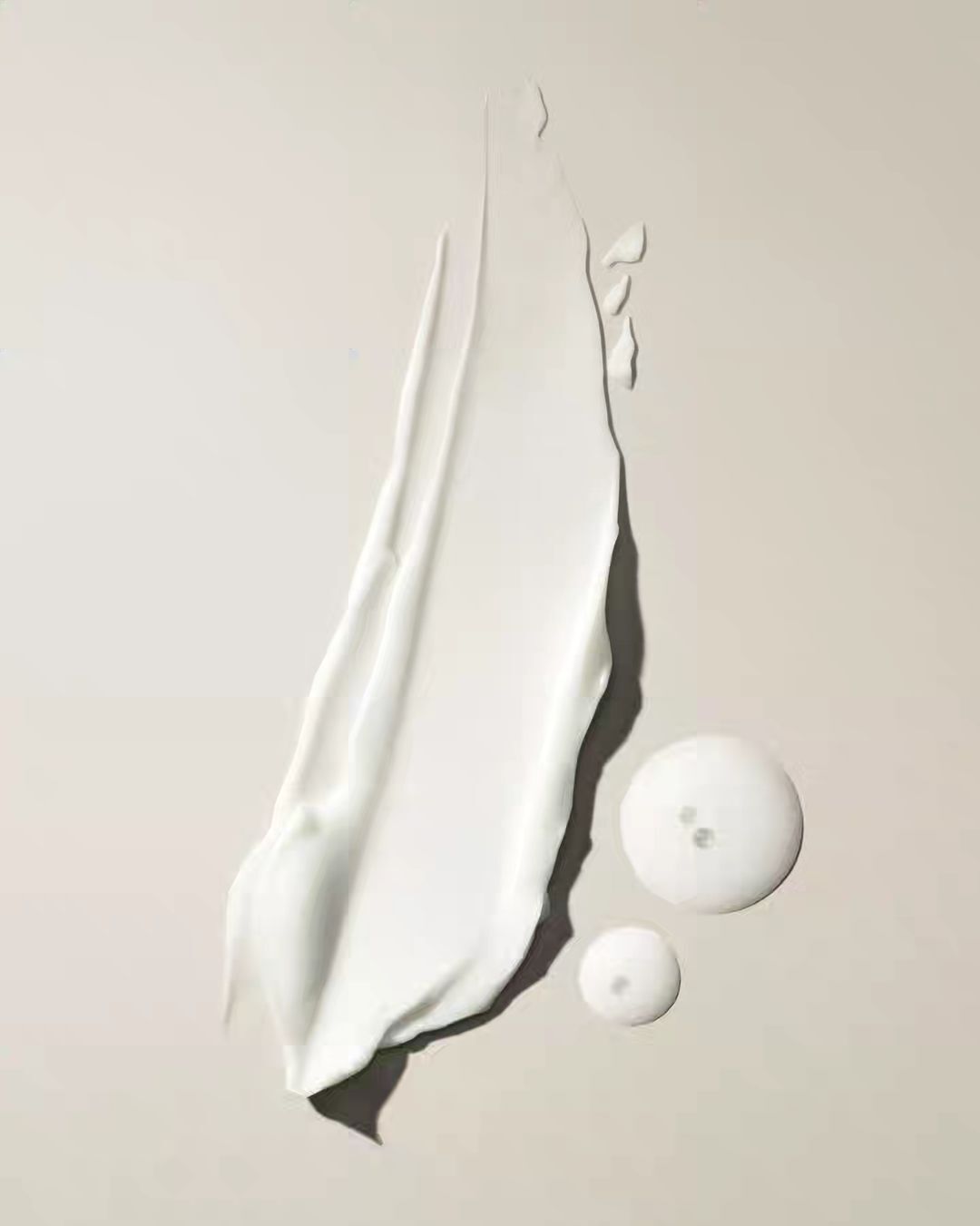 Close-up of a smear of thick, white cream next to two clear gel drops on a neutral surface, studio lit for a beauty product shot.
