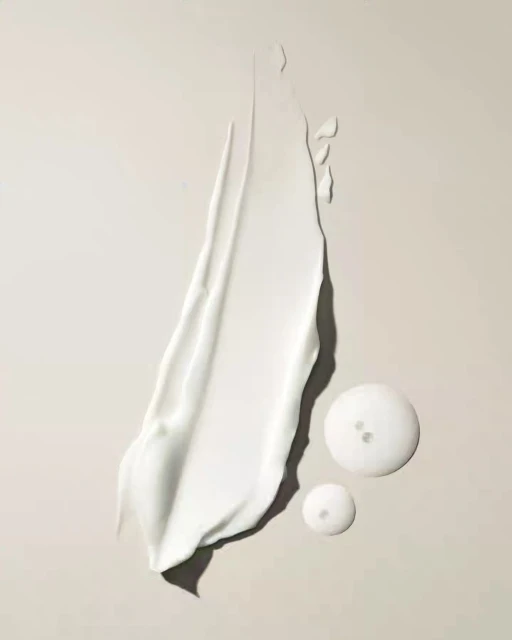 Close-up of a smear of thick, white cream next to two clear gel drops on a neutral surface, studio lit for a beauty product shot.