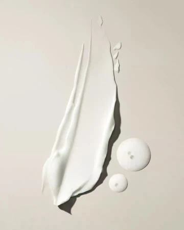 Close-up of a smear of thick, white cream next to two clear gel drops on a neutral surface, studio lit for a beauty product shot.