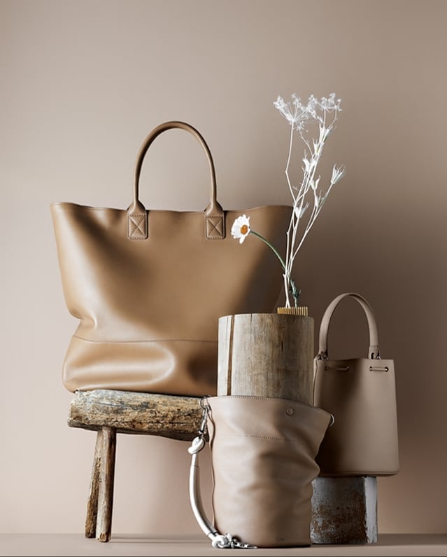 Studio shot of beige leather handbags arranged on rustic wooden surfaces with a single white daisy and ornamental twig arrangement.