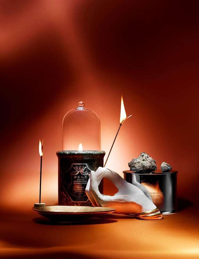Elegant product still life featuring a hand sculpture holding a lit matchstick alongside candles, and containers in warm lighting.