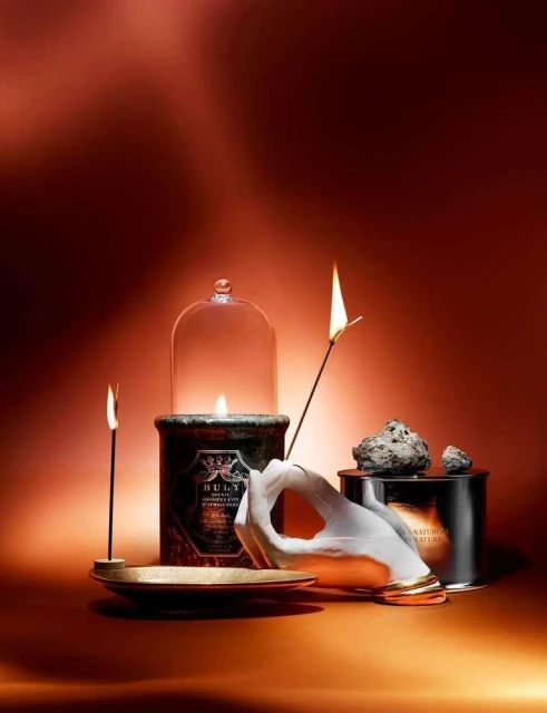 Elegant product still life featuring a hand sculpture holding a lit matchstick alongside candles, and containers in warm lighting.