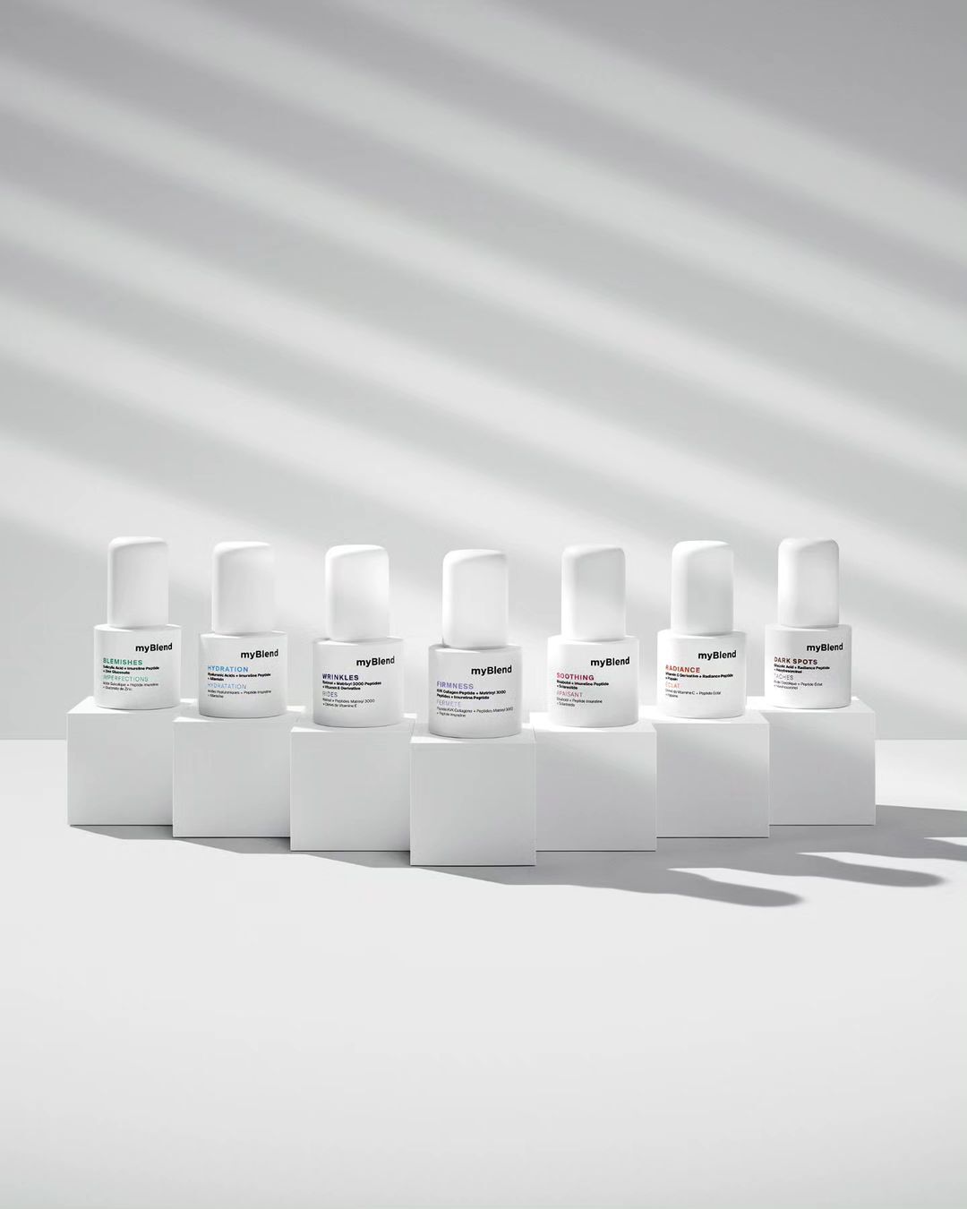 Seven bottles of MyBlend skincare products are arranged on white blocks against a white background with cast window-blind shadows.