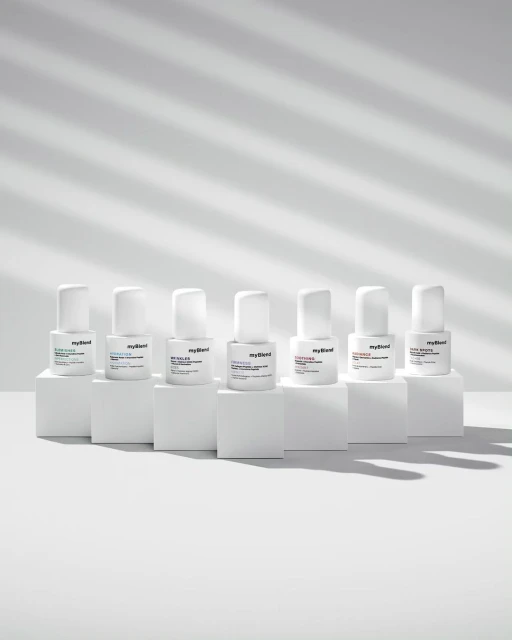 Seven bottles of MyBlend skincare products are arranged on white blocks against a white background with cast window-blind shadows.