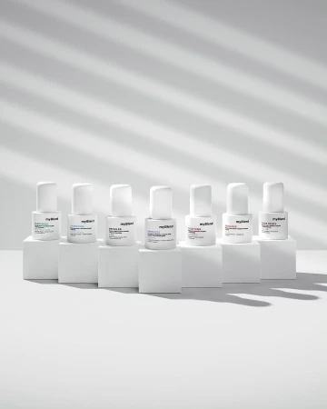 Seven bottles of MyBlend skincare products are arranged on white blocks against a white background with cast window-blind shadows.