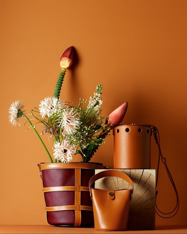 Still life studio shot of designer bucket bags in brown and burgundy leather paired with flowers against a solid tan background.