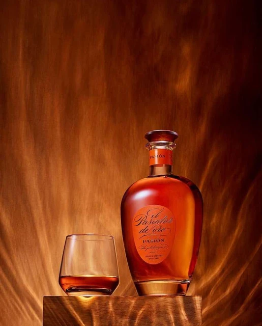 Bottle of El Pasado de Oro rum next to a filled glass, set on a wooden block against a textured golden brown backdrop.