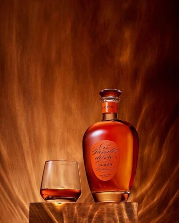 Bottle of El Pasado de Oro rum next to a filled glass, set on a wooden block against a textured golden brown backdrop.