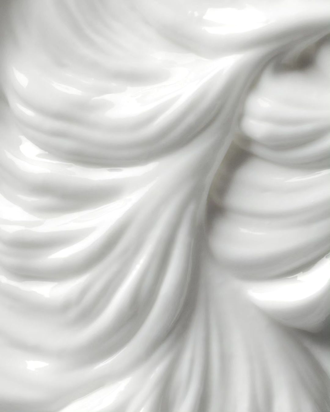 Close-up of glossy white cream with fluid, wavy texture forming smooth peaks and valleys, creating a visually appealing surface.