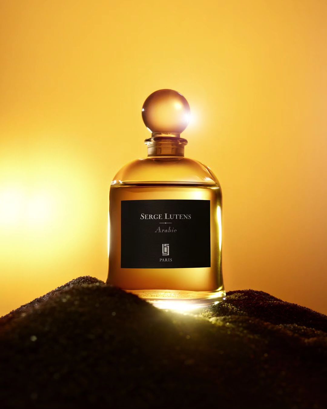 Close-up product shot of a gold-colored perfume bottle with a black label on a small mound of black granules against a gold background.