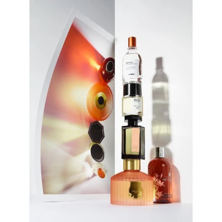 Balanced stack of perfume bottles and jars against a white background with a vibrant color photograph poster on the left