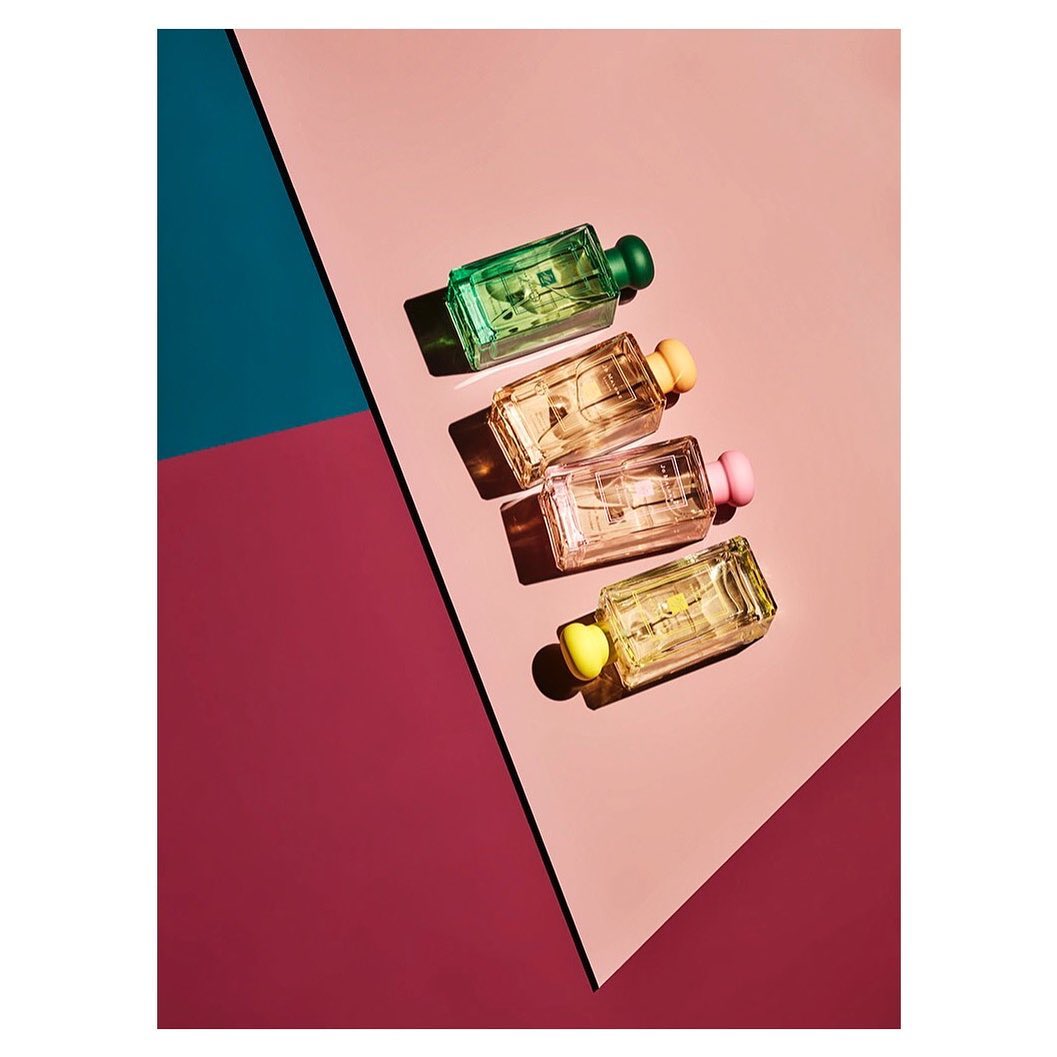Four perfume bottles are displayed diagonally on a pink and red backdrop, with a blue corner, creating a vibrant and eye-catching composition.