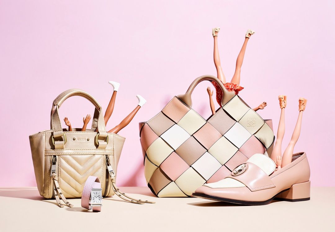 Fashion still life with handbags, a watch, a shoe, and doll legs in a creative composition against a pink background, studio lit.