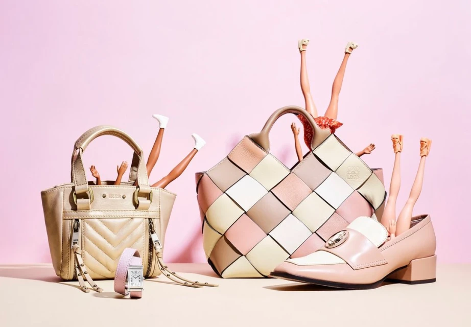 Fashion still life with handbags, a watch, a shoe, and doll legs in a creative composition against a pink background, studio lit.