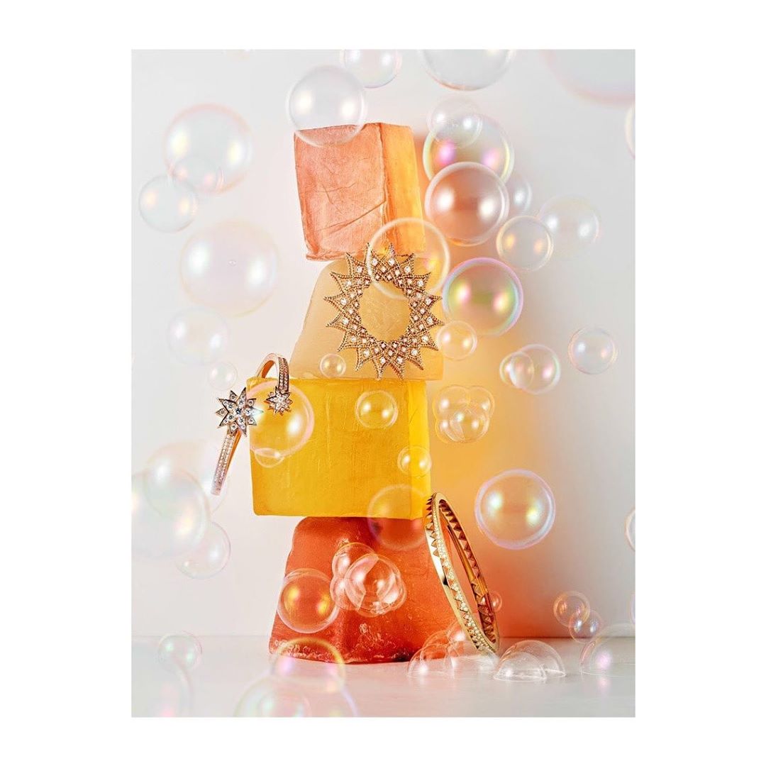 Still life with tower of orange soaps and floating iridescent soap bubbles, gold jewelry and sunlight against white background.
