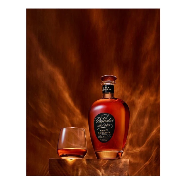 A bottle of El Pasador de Oro Gran Reserva rum sits next to a glass of rum on a wooden surface against a textured copper-toned backdrop.