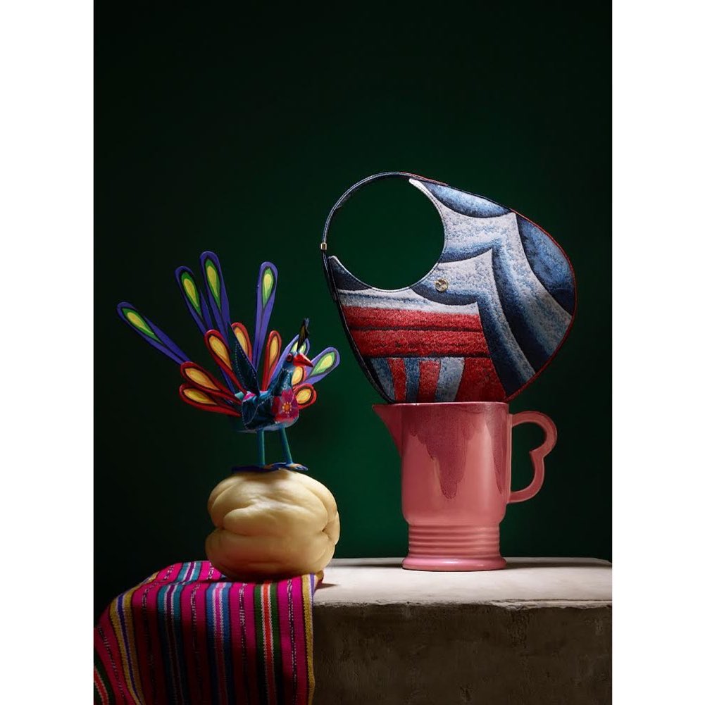 Stylized still life with a designer handbag balanced on a pink ceramic pitcher beside a handcrafted peacock figurine.