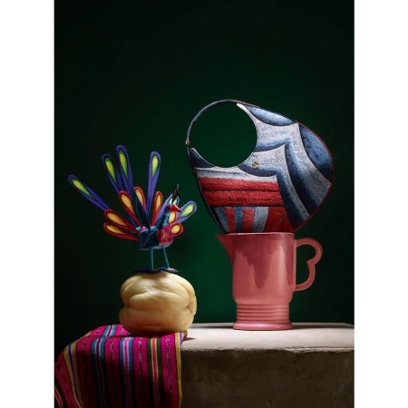 Stylized still life with a designer handbag balanced on a pink ceramic pitcher beside a handcrafted peacock figurine.