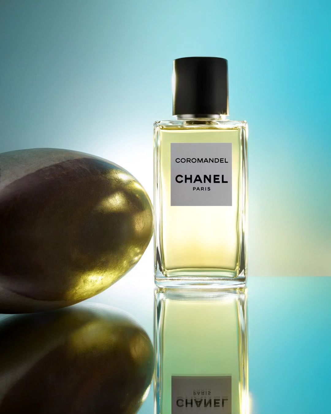 Chanel Coromandel perfume bottle standing next to a smooth golden stone on a reflective cyan gradient surface