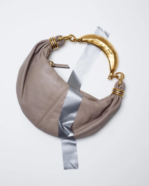 Taupe leather hobo bag with gold handle and silver duct tape on a white surface for fashion or editorial use. Neutral colors.
