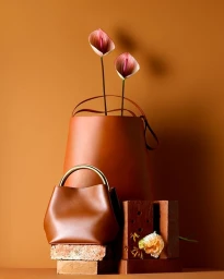 Brown leather handbag with gold handle, tall leather bucket bag holding two anthurium flowers, and bricks on a warm brown background