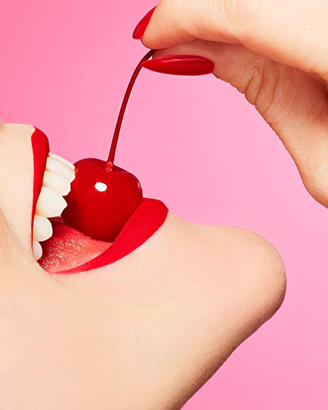 Close-up of a woman's mouth with red lipstick biting a cherry held by a manicured hand against a solid pink background.