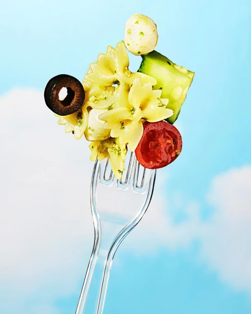 Fork holds bow tie pasta salad with tomatoes, mozzarella, olives, and cucumber against a blue sky background with clouds.