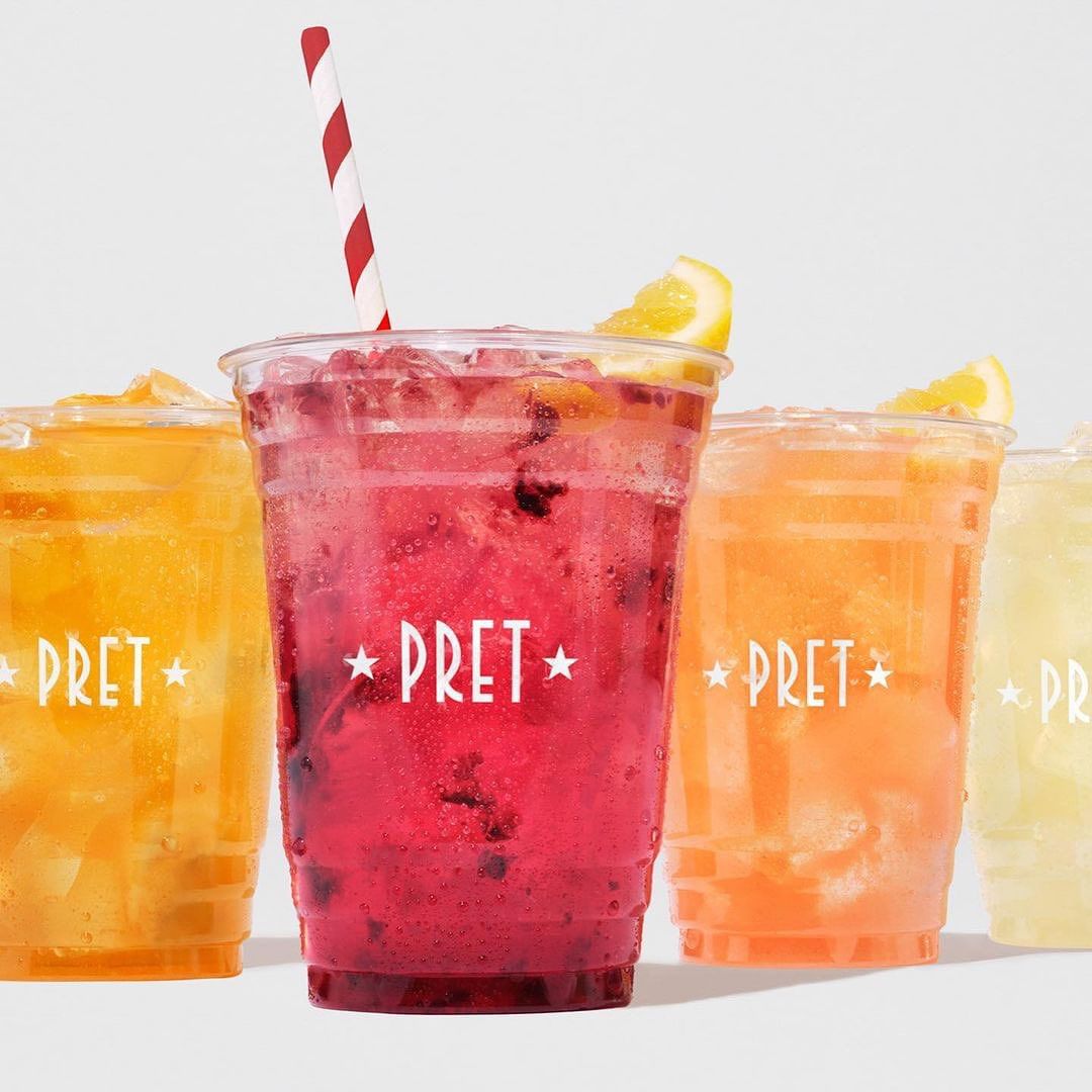 Variety of Pret a Manger fruit coolers in clear plastic cups with striped straws, set against a clean, bright white background.