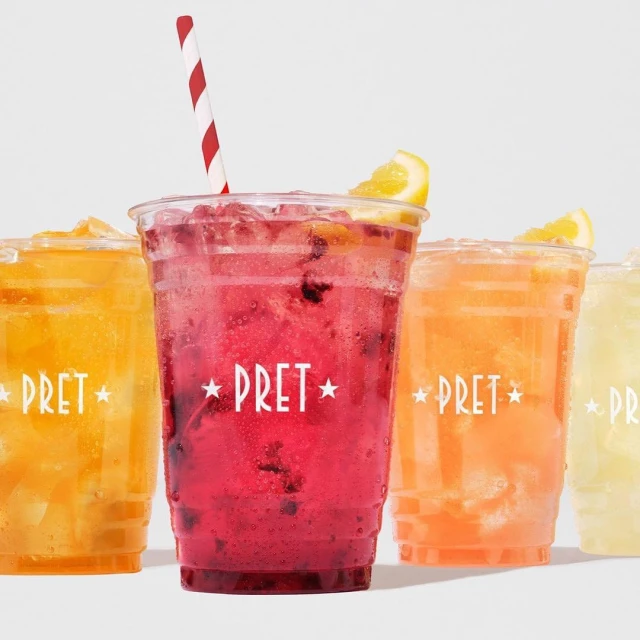 Variety of Pret a Manger fruit coolers in clear plastic cups with striped straws, set against a clean, bright white background.