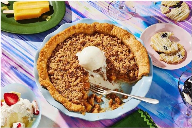 Overhead shot of apple crumble pie topped with vanilla ice cream alongside cookies, sliced strawberries, and a yellow popsicle.