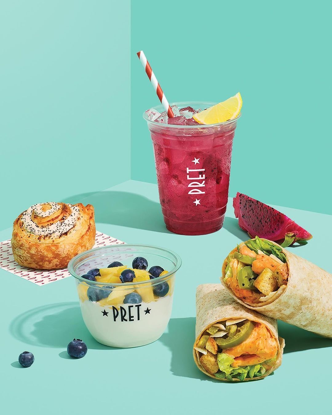 Food and beverage still life features a pastry, yogurt parfait, pink drink, and two wraps, arranged on a light blue surface.
