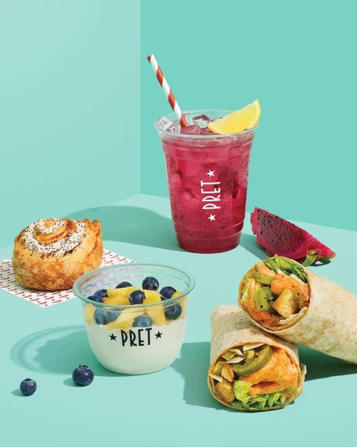 Food and beverage still life features a pastry, yogurt parfait, pink drink, and two wraps, arranged on a light blue surface.