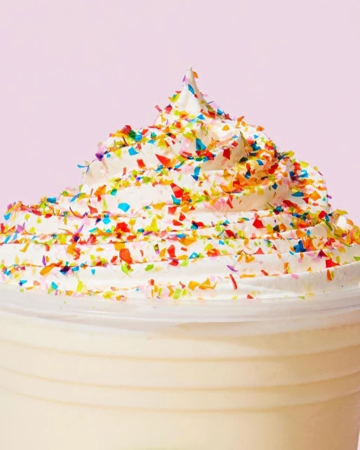 Close-up of a festive milkshake or dessert drink topped with colorful sprinkles on a pink background, showcasing its creamy texture.