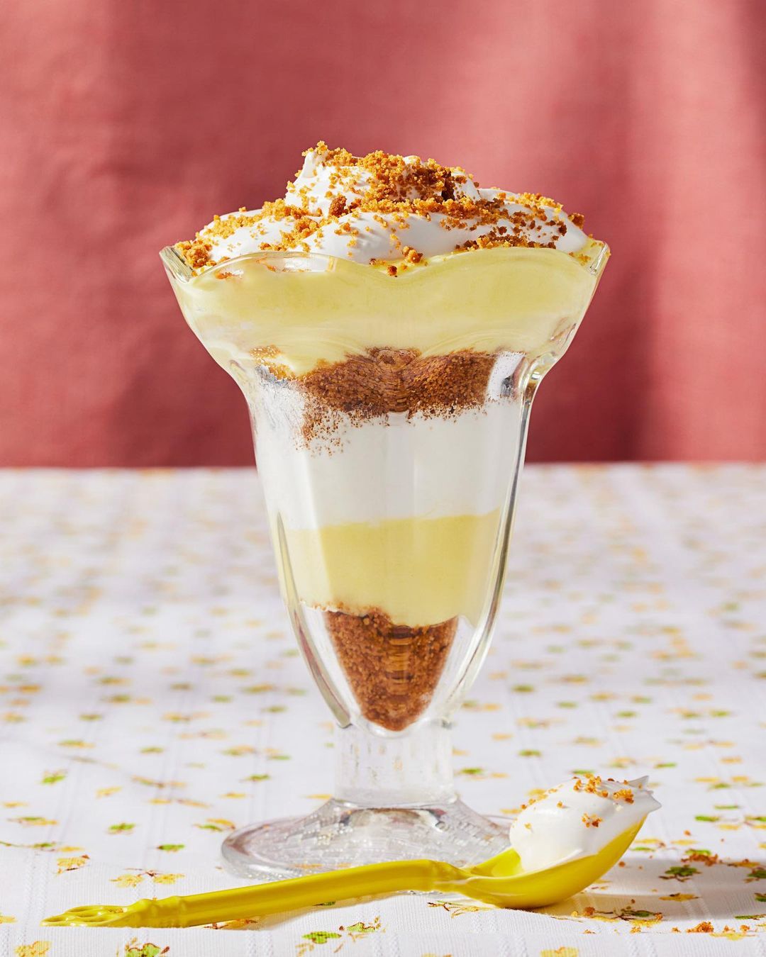 Layered dessert in a glass with whipped cream, custard, and biscuit crumble, standing on a floral tablecloth against a coral background.