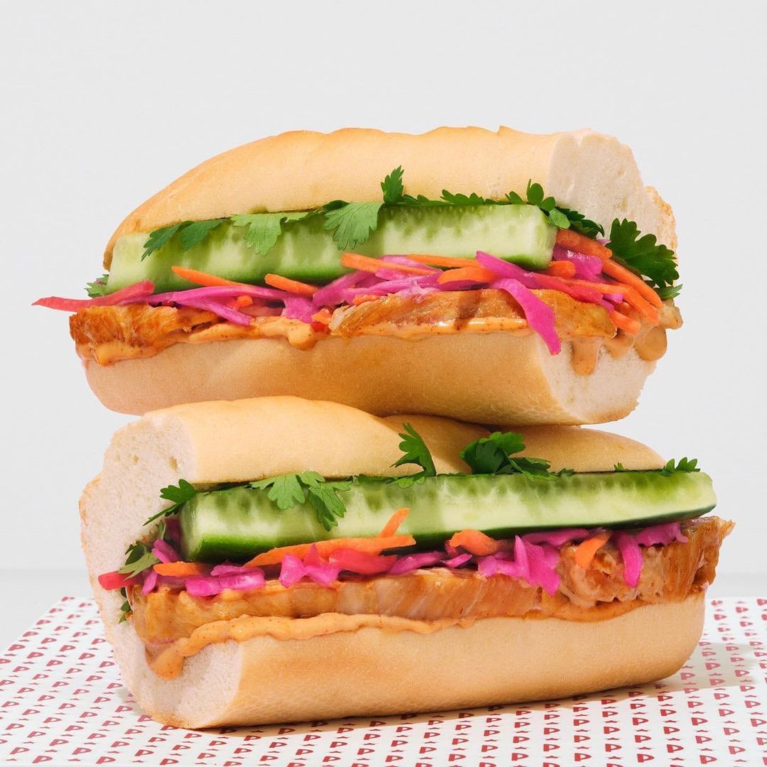 Two appetizing stacked sandwiches featuring fresh cucumber slices, cilantro, and red cabbage on a white surface with a patterned background.