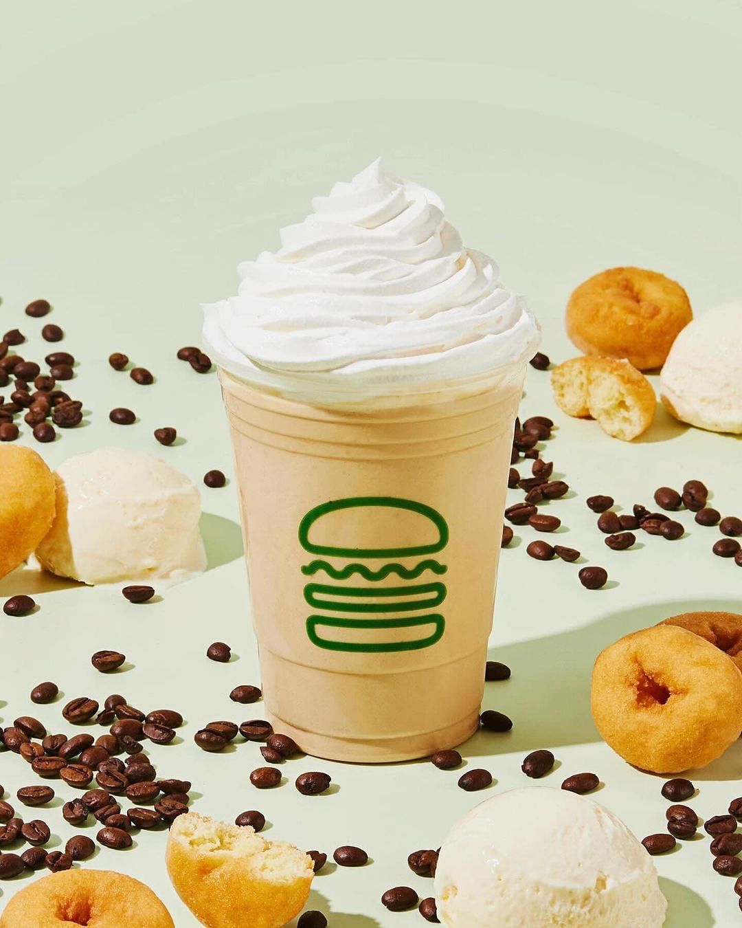 Creamy shake topped with whipped cream, surrounded by coffee beans, donuts, and ice cream against a light green backdrop.