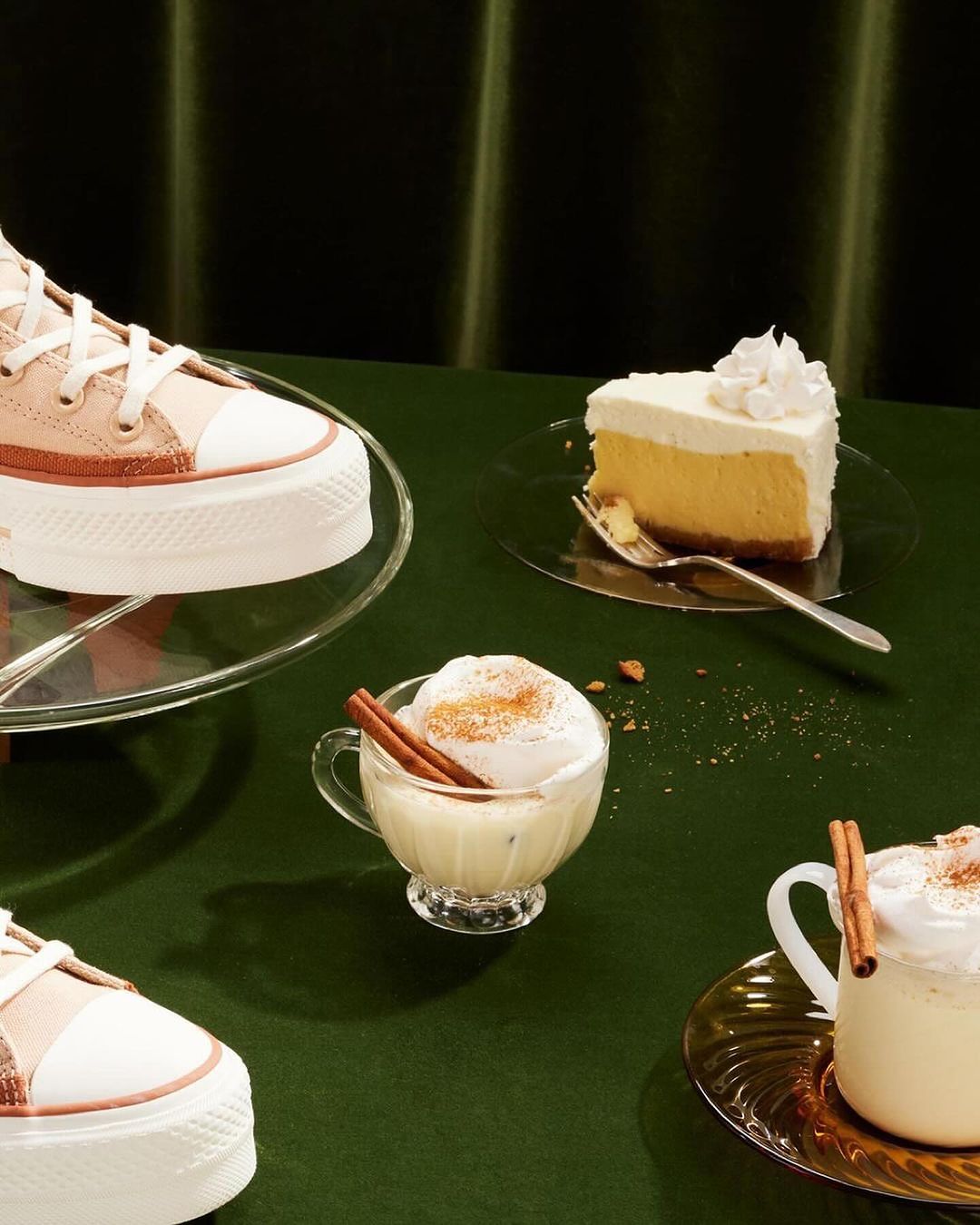 High-angle studio shot: Platform Converse sneakers with eggnog in a glass cup, slice of cheesecake, cinnamon sticks and a green backdrop.