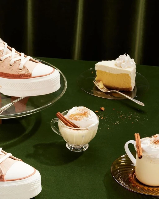 High-angle studio shot: Platform Converse sneakers with eggnog in a glass cup, slice of cheesecake, cinnamon sticks and a green backdrop.