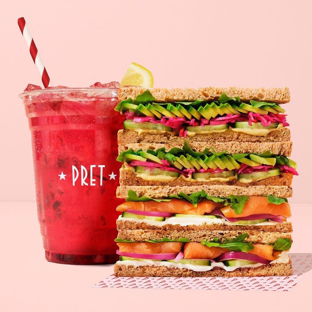 Tall sandwich stacked with smoked salmon, avocado, cucumber, and pink pickled onions, served with a vibrant berry drink with a straw.